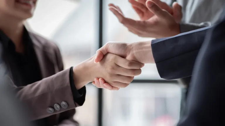 Two business professionals in dark suits shaking hands in an office setting.