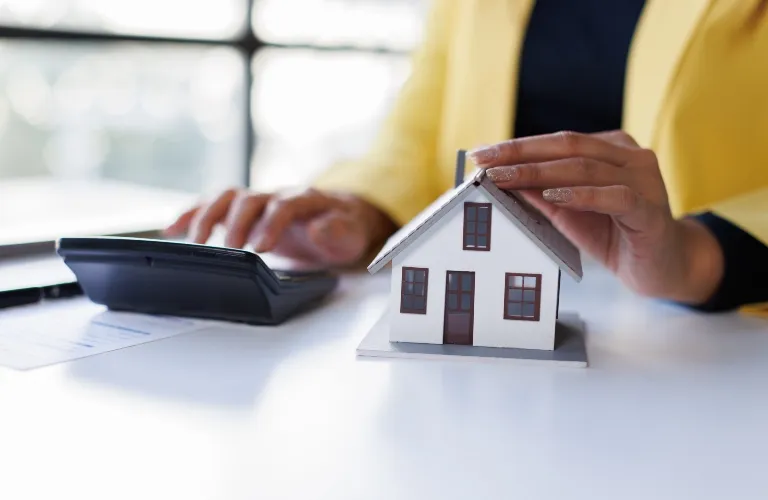 Person in yellow jacket using calculator with small white model house on desk.