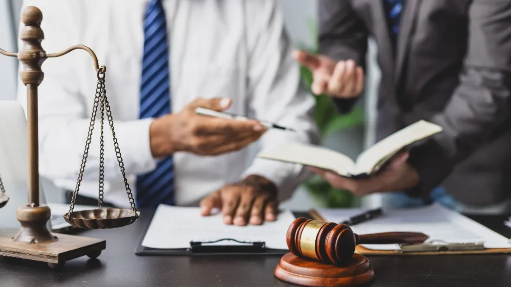 Two professionals in formal attire discussing legal documents with scales of justice and a gavel on the table.
