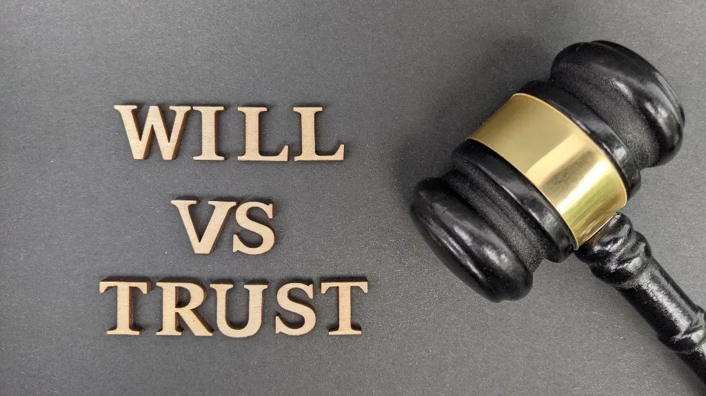 Wooden letters spelling "WILL VS TRUST" beside a black and gold judge's gavel on gray surface.