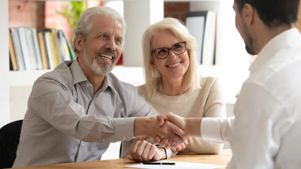 Elderly couple smiling and shaking hands with a professional man across a desk.