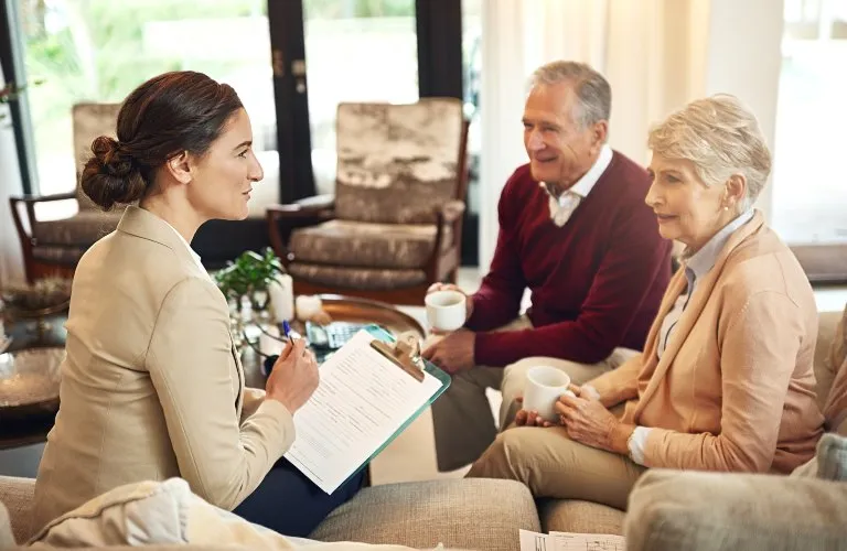 Professional woman with clipboard converses with elderly couple holding white mugs in living room.