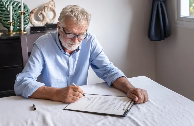 Elderly man with glasses signing a document at a white table indoors.