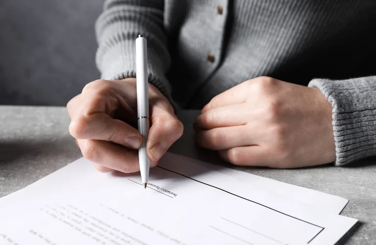 Person wearing a gray sweater signing a document with a silver pen on a gray surface.