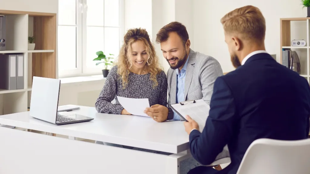 Three professionals in business attire discussing documents at a white office desk.