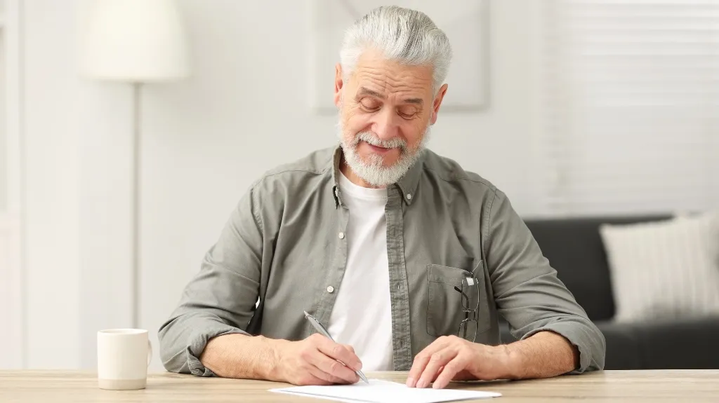 Elderly man with gray hair and beard writing on paper at wooden table indoors.