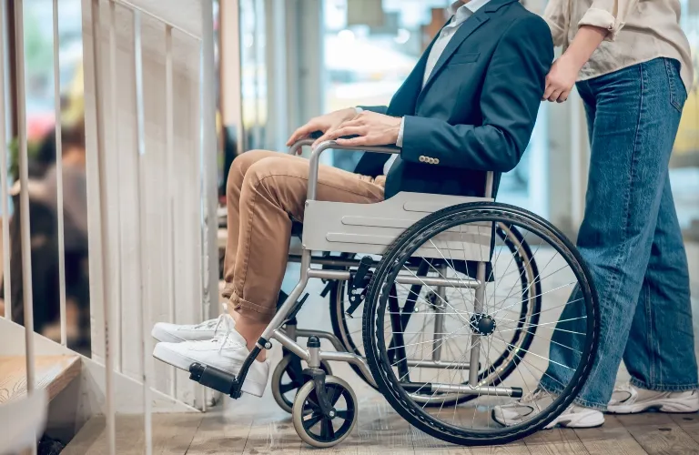 Man in a navy blazer seated in a wheelchair being assisted by a person in casual clothing.
