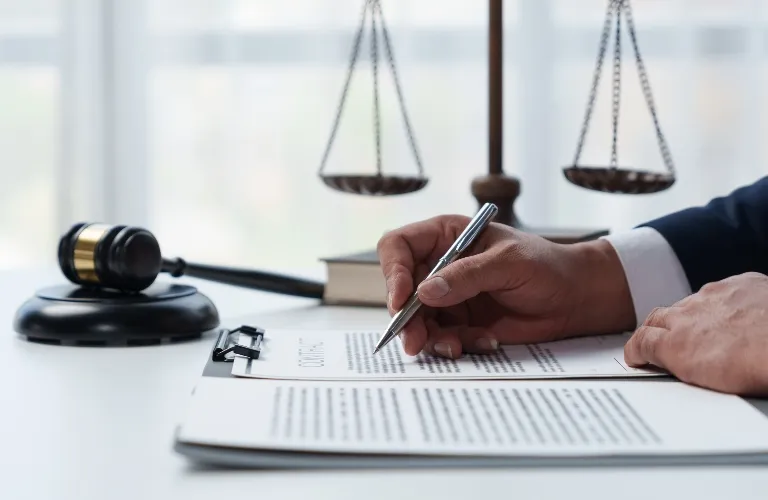 Hand holding pen over legal documents on desk with gavel and scales of justice.