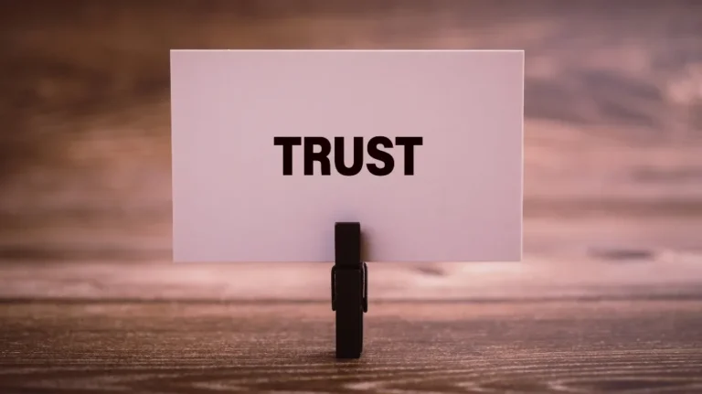 White card with the word "TRUST" in bold black letters clipped on a black stand on a wooden surface.