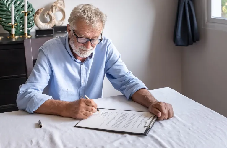 Elderly man with glasses signing a document on a clipboard at a white table.
